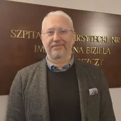 Mężczyzna w okularach i marynarce stoi przed tablicą z napisem "SZPITAL UNIWERSYTECKI NR 2 IM. JANA BIZIELA BYDGOSZCZ".
